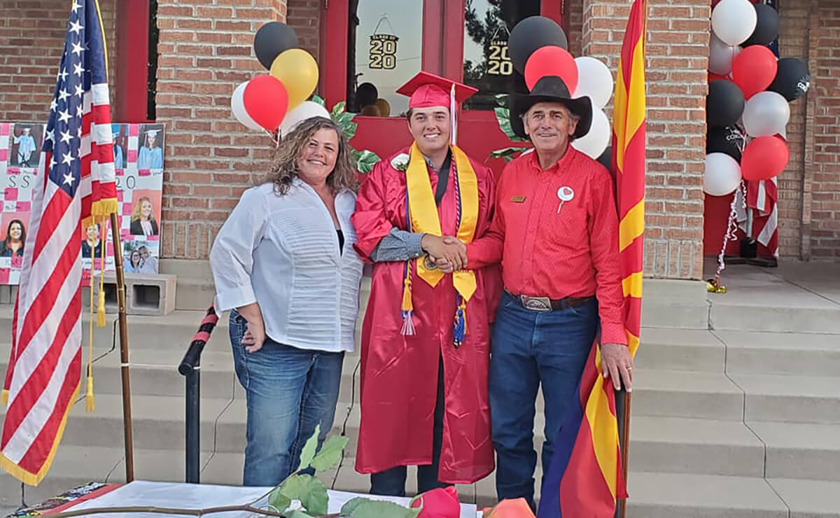 boy wearing graduation cap and gown standing in between parents on school steps