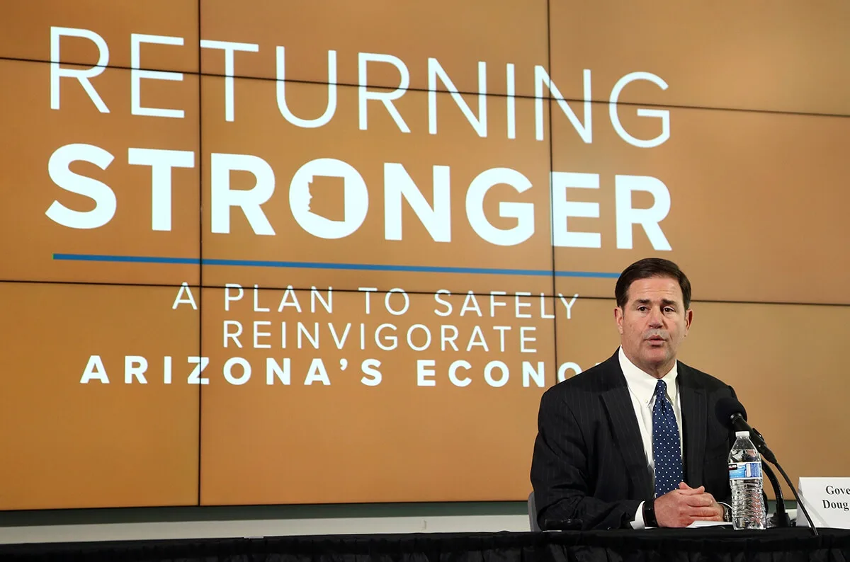 Ducey sitting in front of sign that says "Returning Stronger"