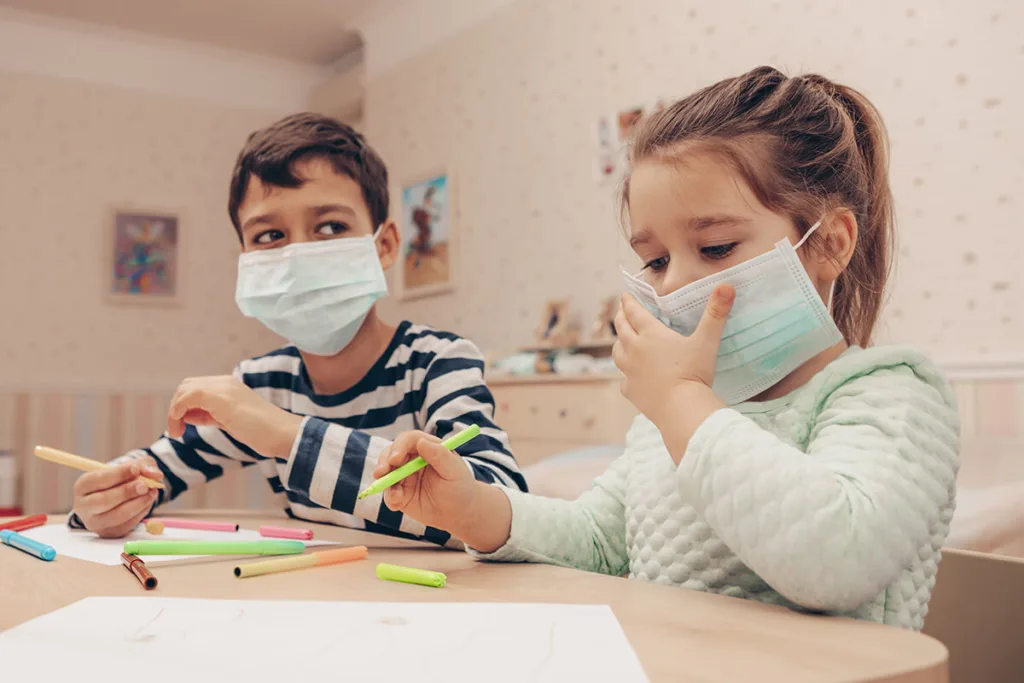 two children wearing face masks sitting at a table drawing