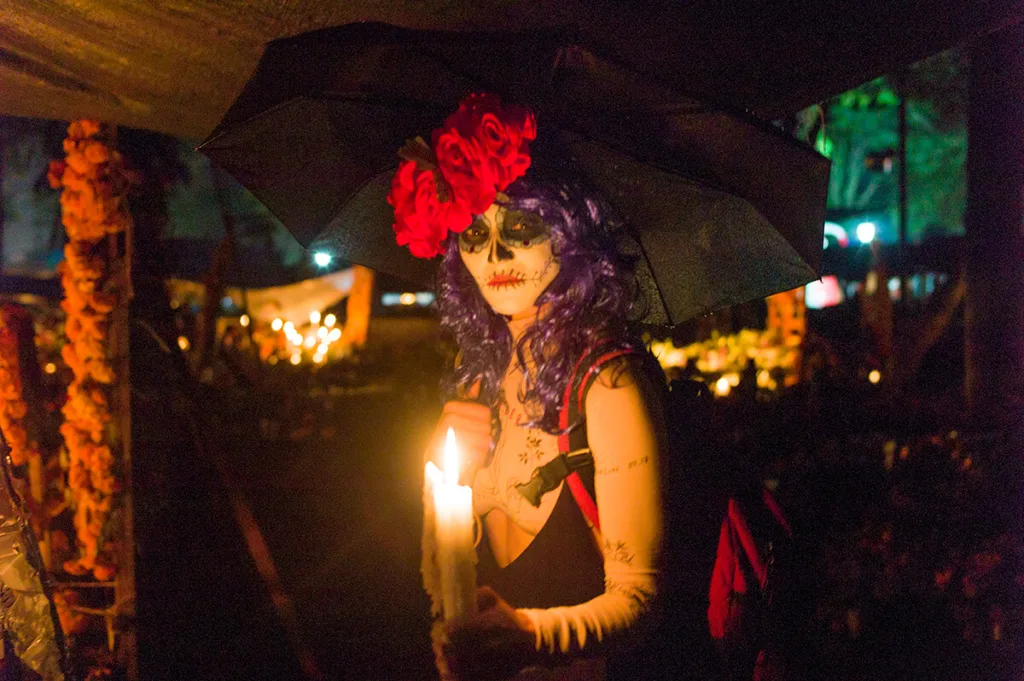 a woman in Day of the Dead makeup holds a candle while posing in the dark