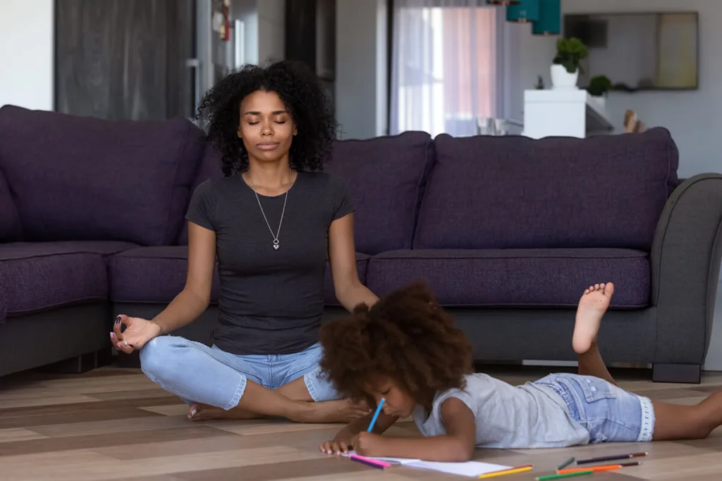 woman sitting on floor meditating while child lays on stomach in front of her coloring