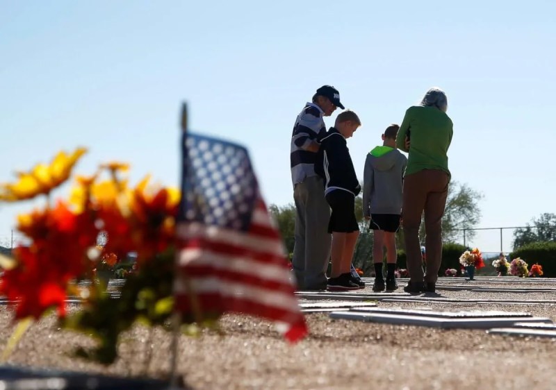 an American flag on a grave in the foreground with a group of people standing over a grave with heads bowed in the background