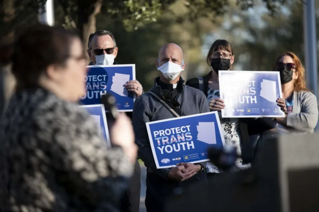 three people holding signs saying "protect trans youth" while woman speaks to crowd