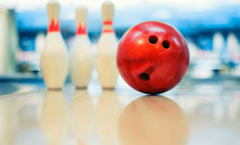 A red bowling ball and three white pins sit on a clean, well-lit bowling lane.