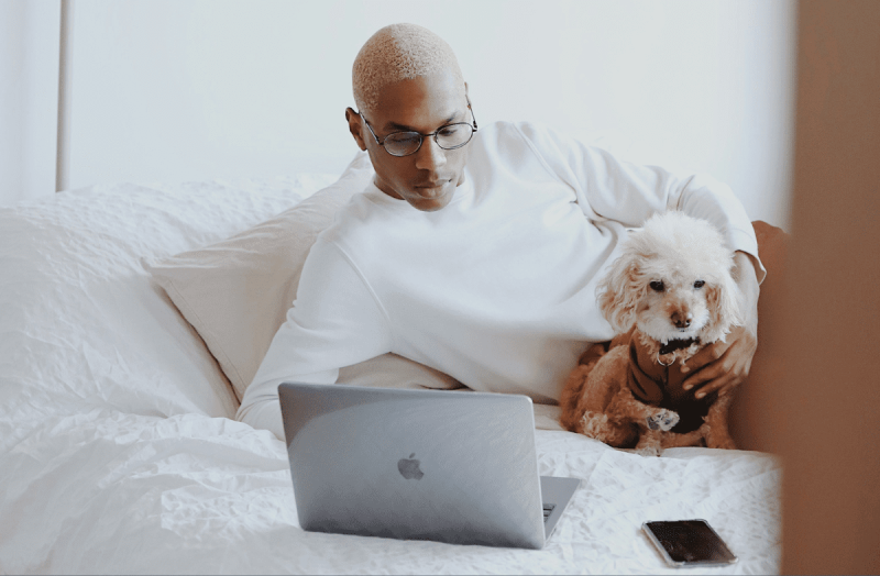 Man sitting on his bed with his small dog and an open laptop.