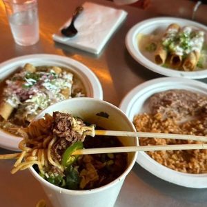 Birria ramen, rolled tacos, and a plate of rice and beans on a wooden table.