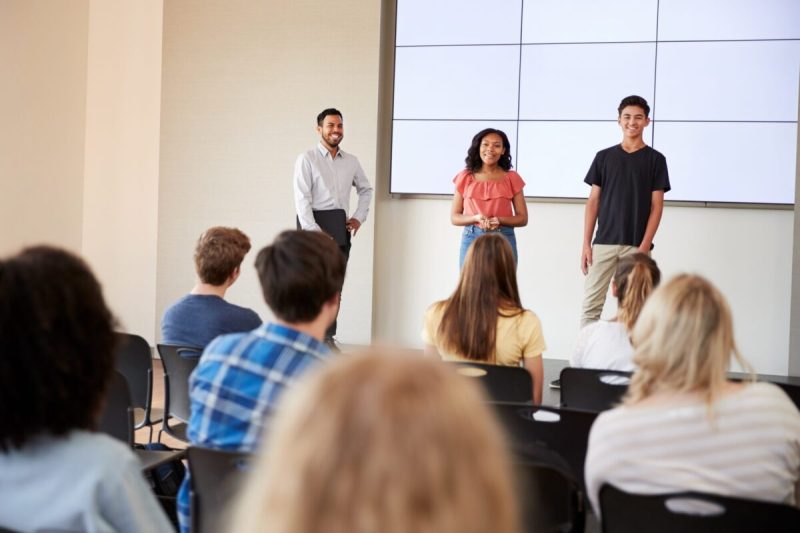 Two Students Giving Presentation To High School Class In Front Of Screen.