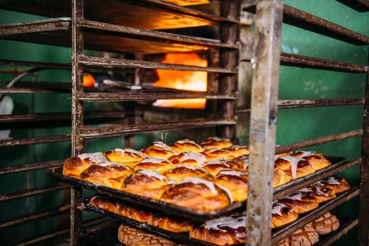 Trays full of sweet bread cooling off beside an oven of a small Mexican bakery.