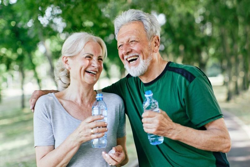 Smiling active senior couple holding water bottles, drinking and jogging together in the park.