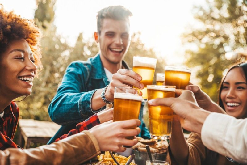 Group of multi ethnic friends having backyard dinner party together - Diverse young people sitting at bar table toasting beer glasses in brewery pub garden - Happy hour, lunch break and youth concept.
