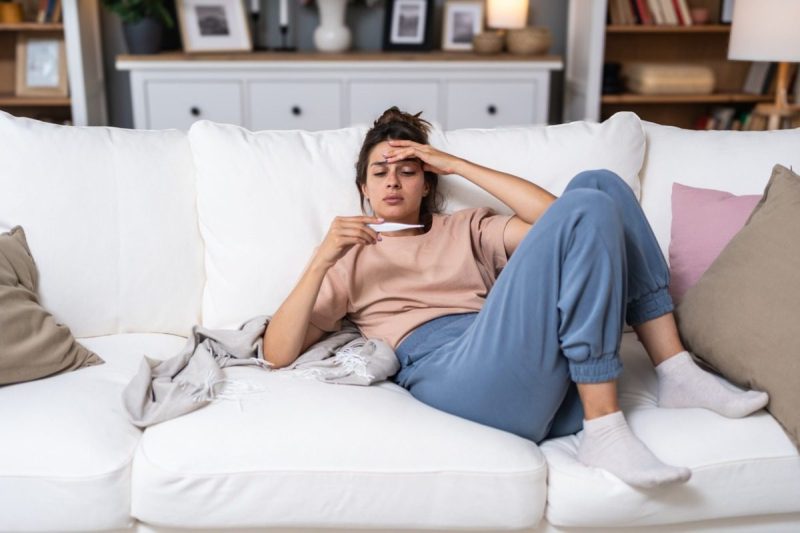Sickness seasonal virus problem concept. Woman being sick having flu lying on sofa holding thermometer. Sick female lying in bed with high fever. Cold flu and migraine with high body temperature.