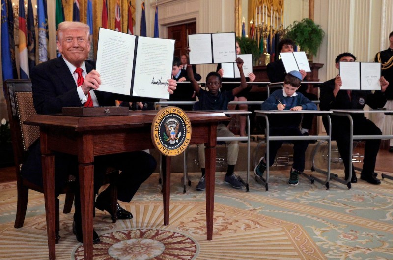 Trump sitting at desk holding up papers alongside children also sitting at desks and holding up papers