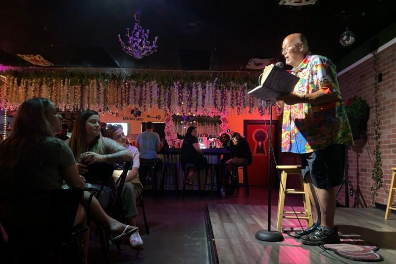 man holding book on stage reading into microphone toward seated audience