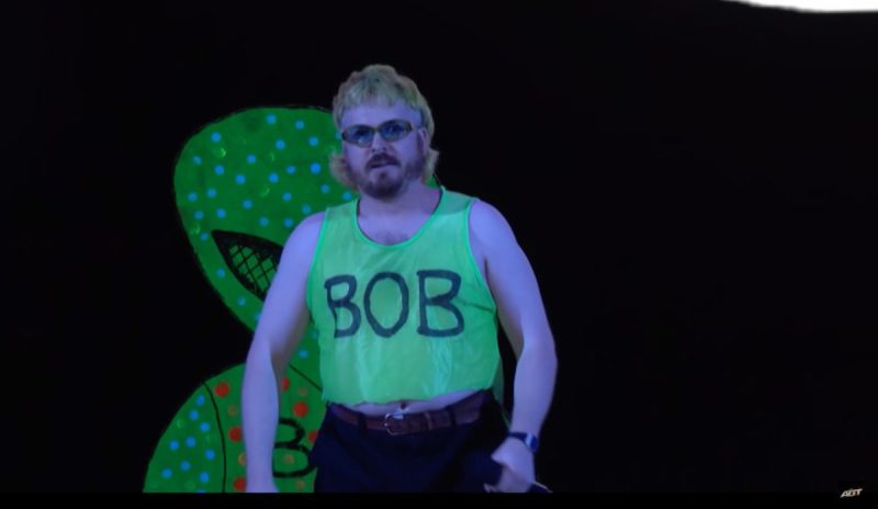 man on stage in silly outfit with alien behind him