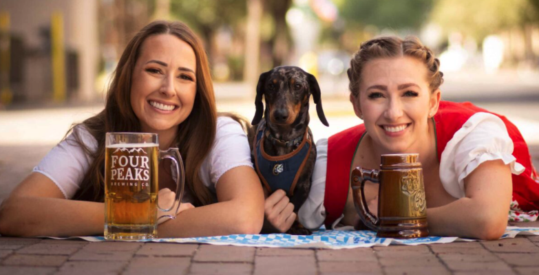 two women in Oktoberfest clothing posing with two beer steins and a dachshund