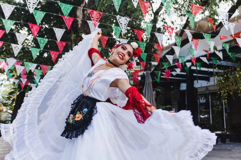 woman wearing traditional Mexican dress dancing in front of flags with the Mexican flag colors