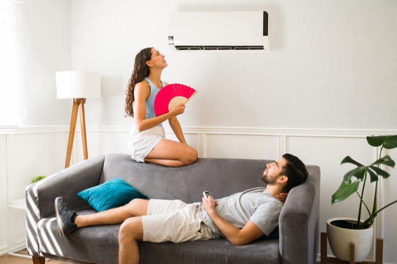 man lying on couch while woman sits on top of couch fanning herself and looking at the AC unit