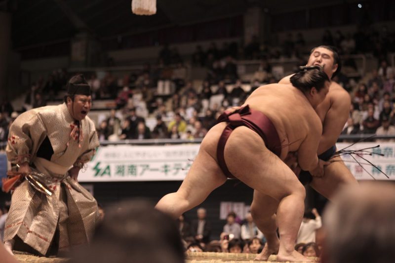 two sumo wrestlers wrestling while referee yells