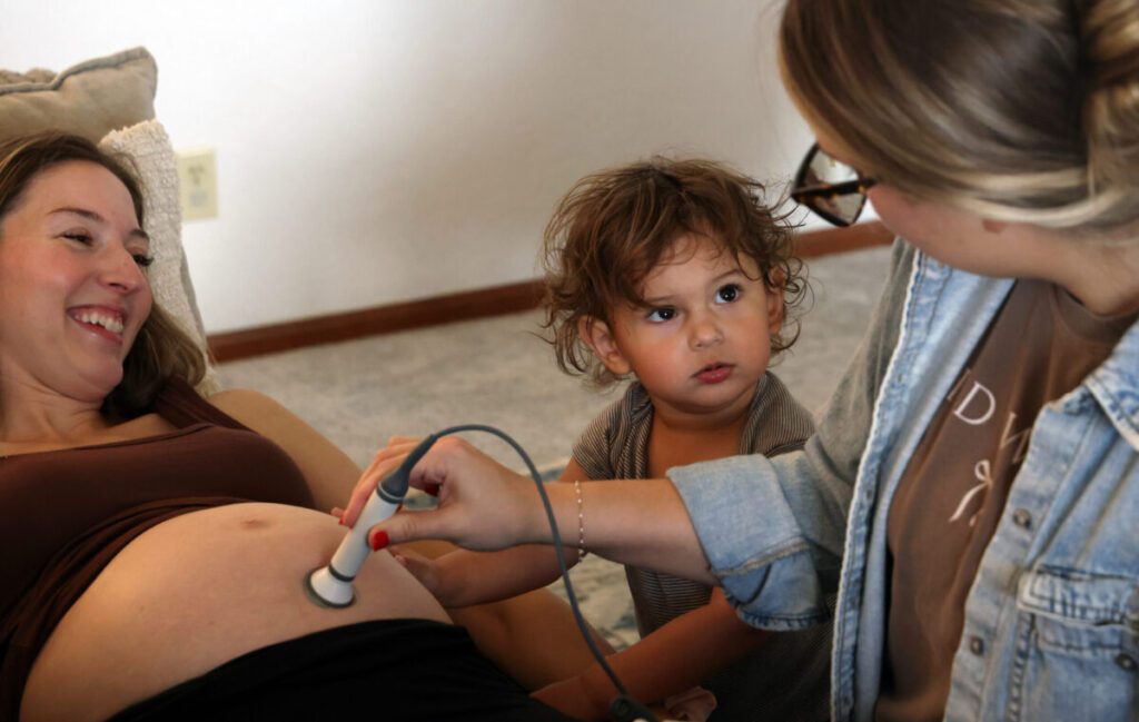 Alberta O’Neill, left, laughs as her son feels her belly during an ultrasound administered by student midwife Nicole Clemens at the O’Neills’ home