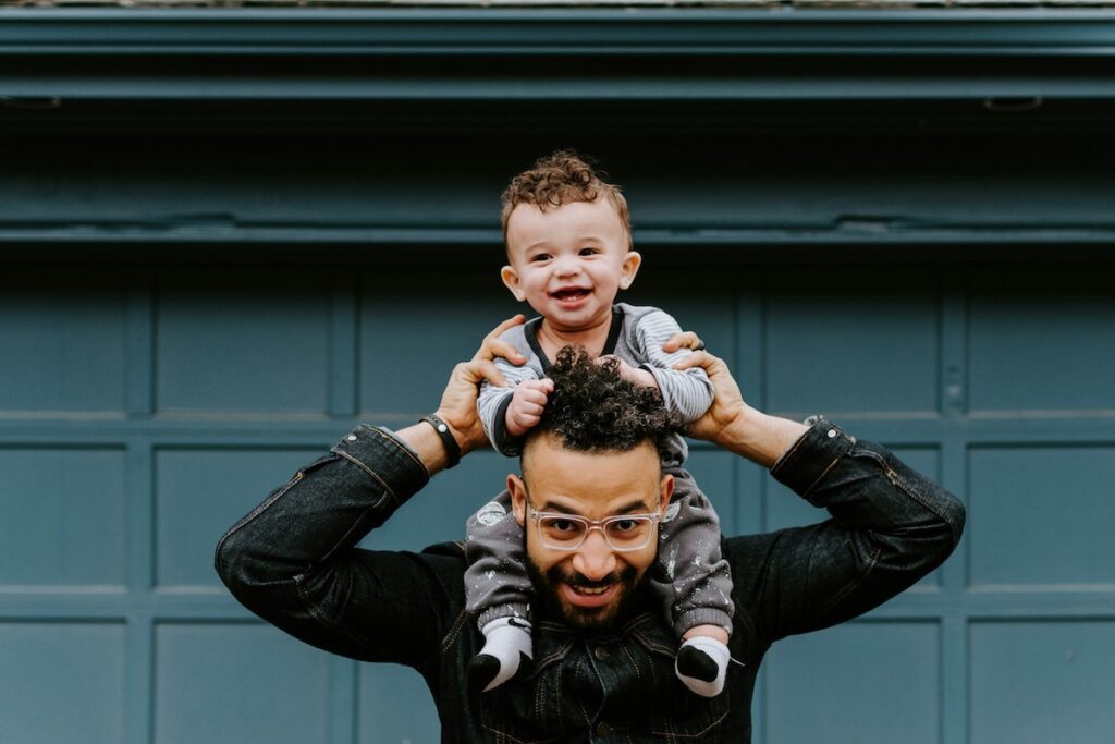 man holding infant on his shoulders in front of a teal garage door