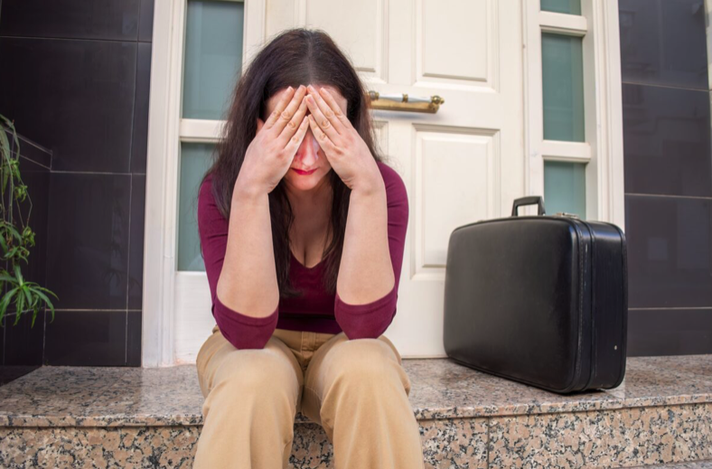 woman sitting on stoop next to suitcase with her head in her hands