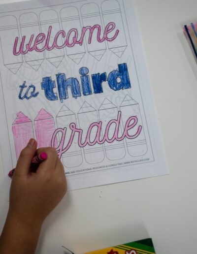 hand coloring in a paper that says "welcome to third grade" on a desk