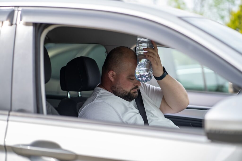 man sits inside car holding water bottle to his forehead while looking down