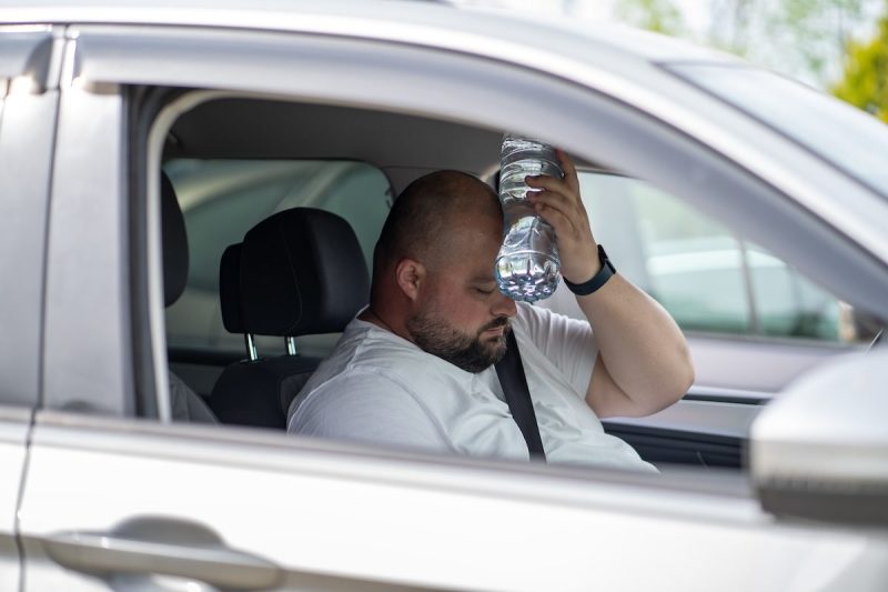 man sits inside car holding water bottle to his forehead while looking down
