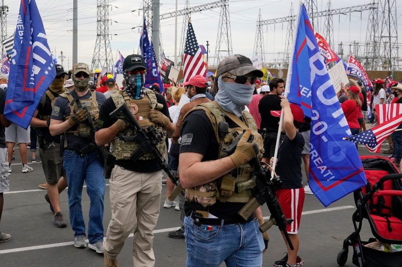 people wearing masks, hats, protective vests, and guns walking in a group holding Trump flags