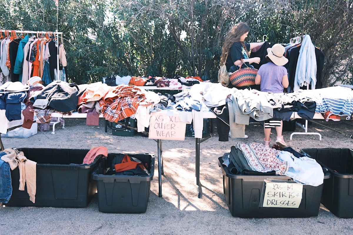 tables with piles of clothes and two people waking through and looking at clothes hanging on a rack