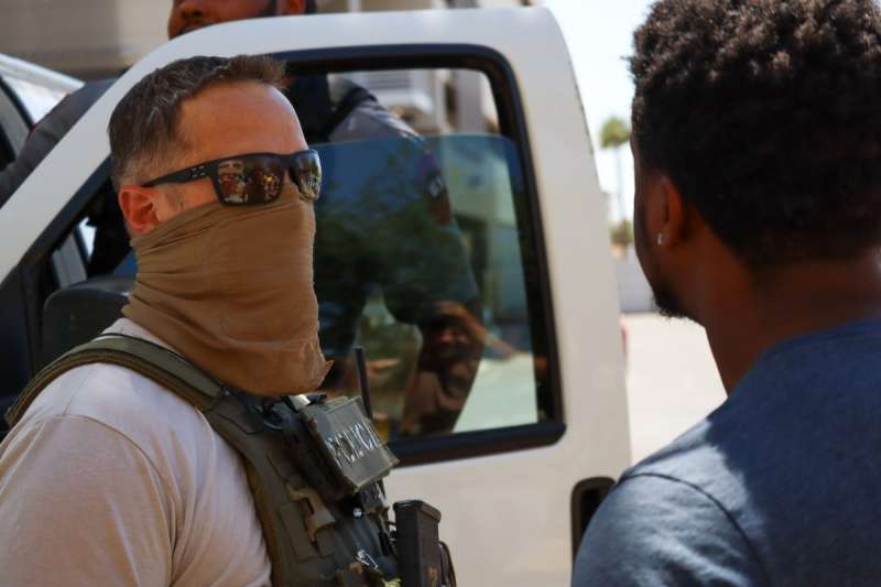 man with bandana on face as a mask and sunglasses standing outside of a truck