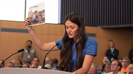 woman holds up a paper while speaking into a mic at a podium
