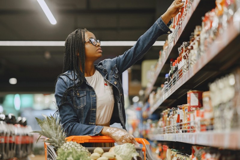 woman reaching from an item on a shelf while pushing a shopping cart filled with groceries