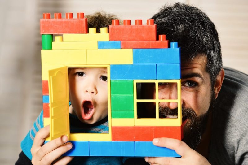 little boy and dad holding up building facade made from jumbo legos as boy makes surprised face in the space of an open door