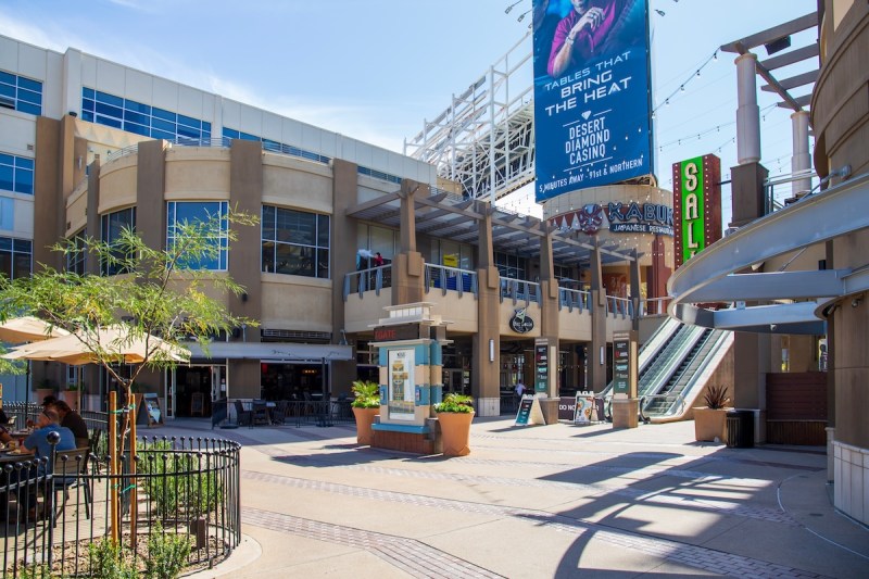 view of stores, restaurants, escalators, and trees in beige shopping outdoor complex