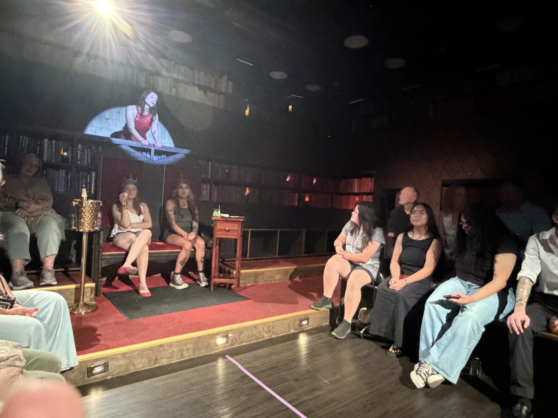 crowd sitting in a theater while a woman stands at a balcony in a spotlight 