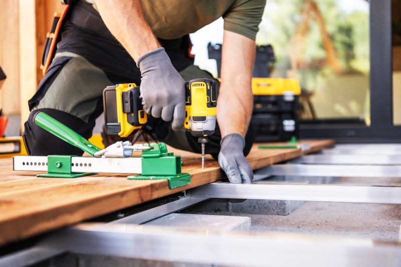 A skilled craftsman uses a drill to secure wooden planks on a deck frame while wearing gloves. Tools and building materials are organized nearby in a home setting.