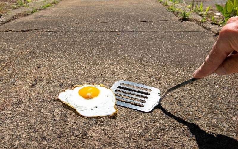 a fried egg on a sidewalk and someone putting a spatula underneath it