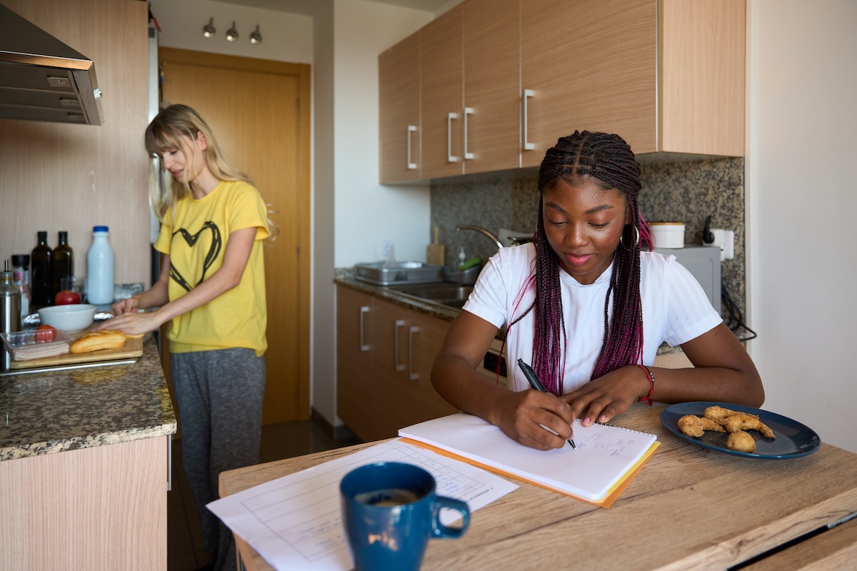 woman standing at kitchen counter preparing food while another woman sits at a table writing in a notebook