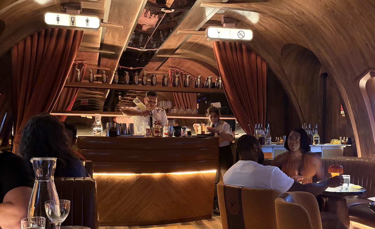 people dressed as flight attendants making drinks at a bar in a room designed to look like a vintage airplane lounge