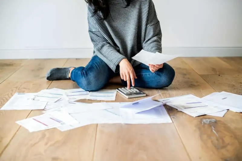 woman sitting on floor with paper bills spread out around her while she pushes a button on a calculator