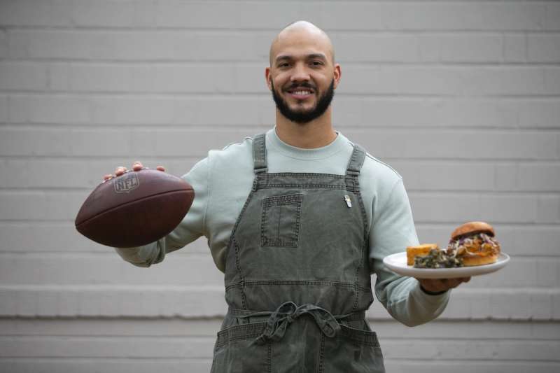 man wearing apron holding football in one hand and plate of food in the other