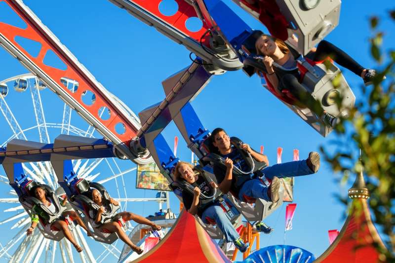 people riding on a spinning ride with a ferris wheel in the background at a state fair